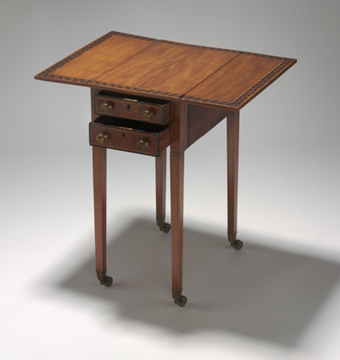 Rectangular wooden table, with fold-down side leaves. The table features narrow inlaid ivory borders, including a wide inlaid border of contrasting timber in a geometric pattern on the table top. The table has four square tapering wooden legs, each terminating in a brass cup and castor. The table has two drawers, each with two round metal knob handles and a brass key-operated lock.