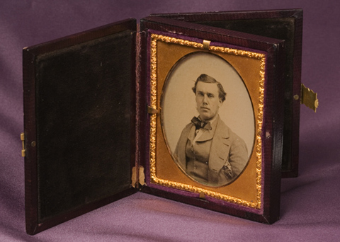 A leather covered case with a metal clasp on one side which opens to reveal an oval glass ambrotype photograph of a man wearing a suit and cravat. The photo is contained within a gold frame. The case is lined on both sides with black velvet which provides a background against which both sides of the photo can be seen. The leather on the case is worn and one side is completely detached from the body of the case.