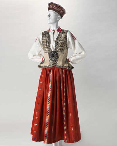 Mannequin dressed in a Latvian black silk bonnet with a drawstring cord at the neck, a red ankle-length skirt, a white linen shirt and a waistcoat decorated with an embroidered black pattern of geometric curves.