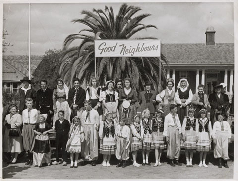 Black and white photograph of a group of adults and children standing in two rows, wearing various traditional national costumes. The group stands beneath a 'Good Neighbours' sign and in front of a palm tree and building.