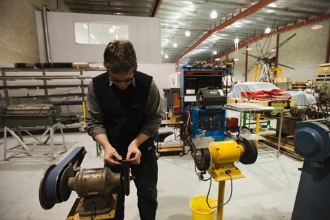 A man wearing safety glasses bends over a machine, holding a small bolt over a wire brush. Various other machines are visible in the background, along with the frame of a large windmill.