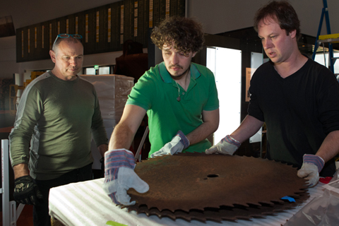 A colour photograph of two men heavy, sharp metal blades. A another man is standing nearby watching them.
