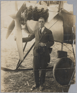 A black and white photo of a man standing at the front of plane, with one hand resting on a propeller blade.