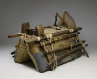 A camel packsaddle with a wooden frame secured with ropes. The wooden frame surrounds padded hessian and cloth bags.