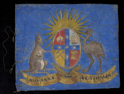 A hand-painted flag depicting a kangaroo and emu on either side of a shield, against a bright blue background. The shield is broken into four sections showing a sheep, a sailing ship, a sheaf of wheat and a pick and shovel. The stars of the Southern Cross separate the quadrants and a rising sun appears above the shield. A banner below the coat of arms reads 'ADVANCE AUSTRALIA'. At the left edge a hoist cord stitched at two points to the flag.