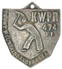 An image of a worker holding a tool above his head and in a digging stance is in the centre of the medal. 'KWPR 474 FT' is written in the top right corner and 'Snowy Mountains Australia' is written down the left hand side.