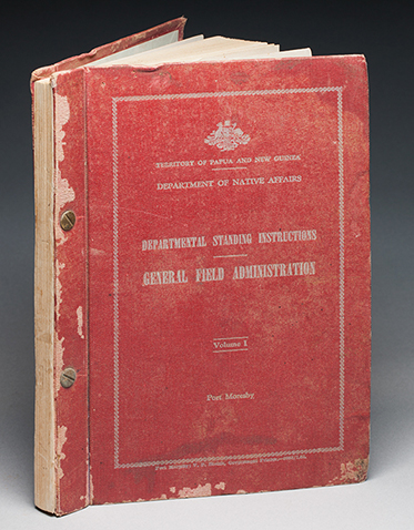 A red hardcover book with a loose leaf inside the front cover. The book features two gold coloured fixings through spine, and silver coloured text on the front cover, which is within a silver border and reads 'TERRITORY OF PAPUA AND NEW GUINEA / DEPARTMENT OF NATIVE AFFAIRS / DEPARTMENTAL STANDING INSTRUCTIONS / GENERAL FIELD ADMINISTRATION / Volume 1 / Port Morseby'.