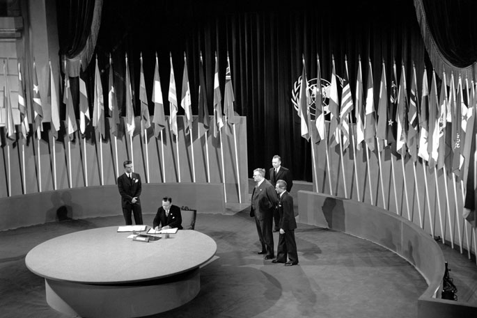 Interior black and white photo of man sitting at large round table signing a document, with four men standing behind him and a series of flags arranged behind them.