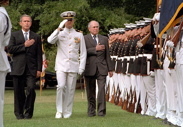 President Bush and Prime Minister Howard walking with hands on hearts on either side of a US Navy officer. A line of marines in full-dress uniform stand to attention on their left.