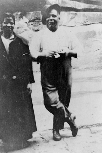 Aboriginal man, smiling, stands facing the camera, a hat set at jaunty angle on his head. An Aboriginal woman stands to his right
