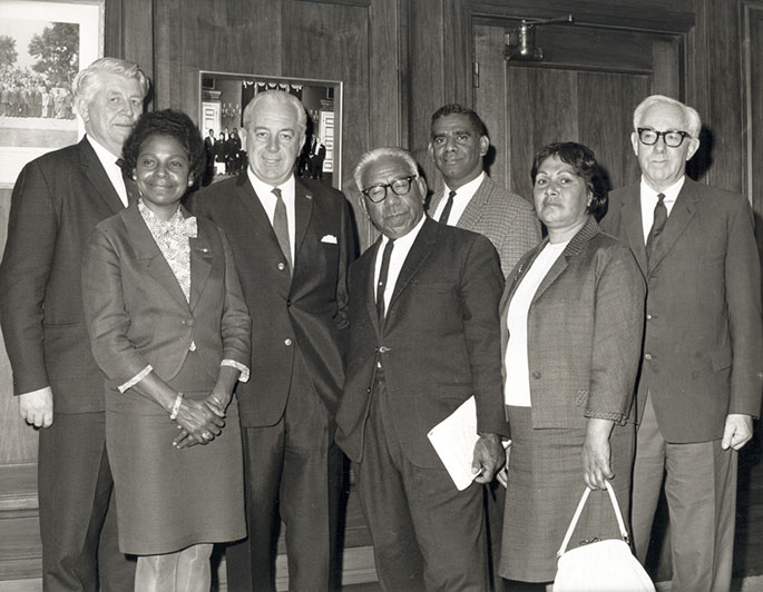 The seven people named stand in front of a wood-panelled wall in what looks like Old Parliament House.