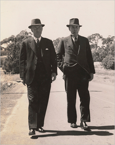 two men in suits and hats walk side by side outside.