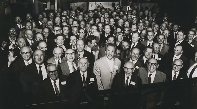 interior photo of tightly packed group of about 70 men in their 50s and 60s smiling at the camera.