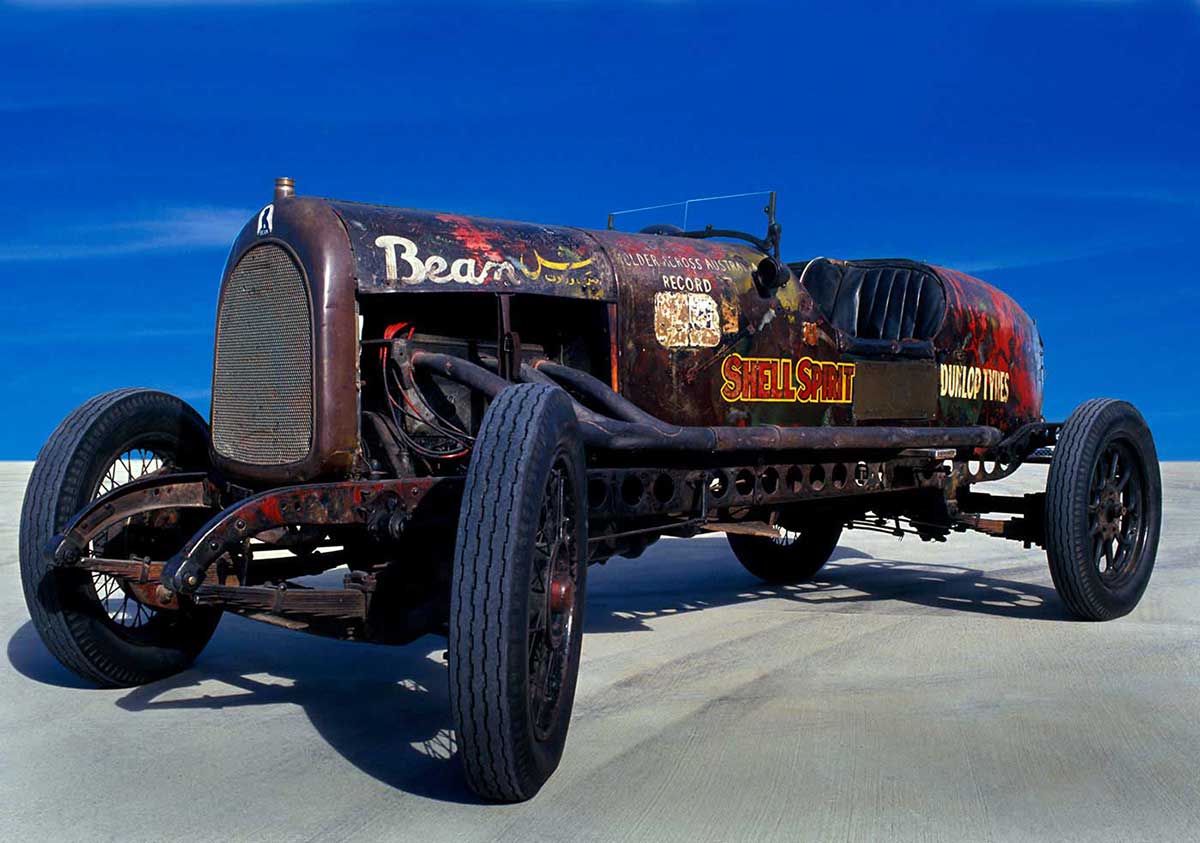 A four-wheeled open seater car with a long engine cowling, boat tail and low glass windscreen. 'Bean' has been painted in white on the top right of the bonnet cover. The words 'Shell Spirit' and 'Dunlop Tyres' are visible on the right side of the car's body.