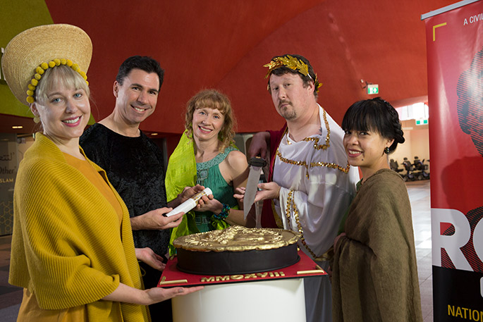 Five people dressed in costume surround a large cake in the form of a Roman coin