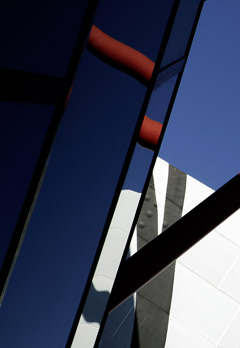 Part of the exterior of a museum. The image shows part of a window that is separated into several sections. Diagonal lines divide up the window; reflections of blue sky and parts of the museum's structure can be seen in each section. The upper left corner of the image is darker than the rest.