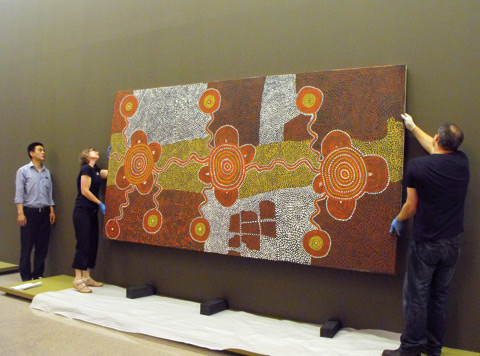 A colour photograph of three museum staff installing a large painting in an exhibition. The painting features traditional patterns in ochres, browns, yellows and white. Two people wearing gloves hold the painting up off the floor. Three support blocks can be seen on the floor underneath the painting. The wall in the background where the painting will be hung is olive green.