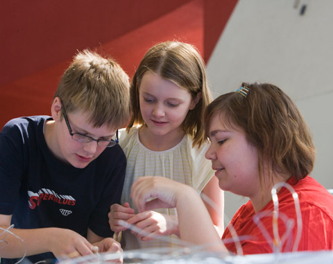 Two young children and a woman intently engaged in a craft activity. Loops of string are out of focus in the foreground; in the background the red and white walls of the Museum's Hall are visible.
