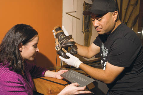 Helen Sartori holds a small text panel while John Atafu, wearing white gloves, holds a wooden canoe prow sculpture.