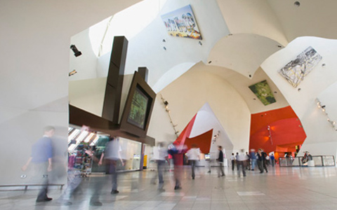 An image of the Museum's Hall with a number of people walking across the large open space, their images blurred and ghost-like. The vaulted white ceiling of the Hall dominates the image with a number of paintings hanging from the ceiling.