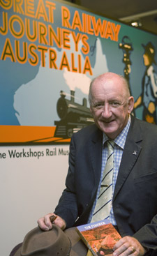 Tim Fischer, sitting holding his hat and and a book, in front of the Great Railway Journeys of Australia exhibition banner.