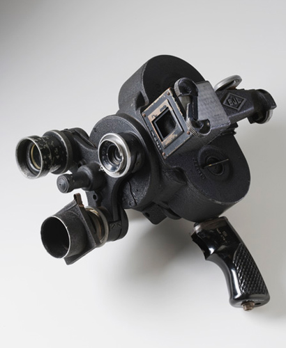 A black-coloured metal motion picture camera with three-lens turret, clockwork-driven film cartridge and side viewfinder. A black pistol-style handgrip extends beneath the camera body.