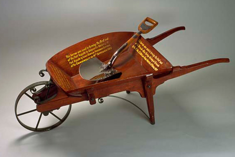 A wooden wheel barrow, with yellow script painted on the inside, and a metal shovel
