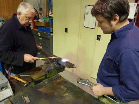 A man applies heat to a piece of metal, watched by another man, who holds a wrench.
