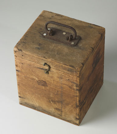 Wooden box with a lid. It has a small metal latch hook at the front and a rusted metal handle on top.