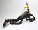 A black and pink model camel and rider made from, raffia, minarri (greybeard) grass, wool and leather. The rider has a yellow and black body, is wearing a purple beanie, and is seated on a brown crocheted saddle which is fastened to the camel with leather straps and leads.