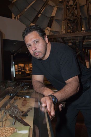 lee-burgess-1.jpg A man leans on a glass display case containing a number of objects. A large windmill can be seen in the background.