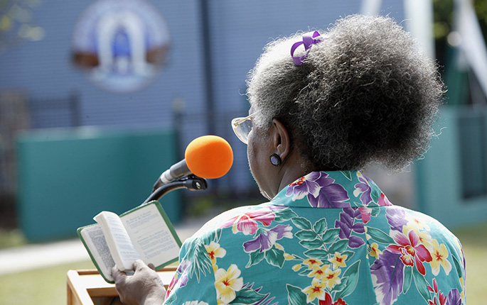 A coloured photograph of the back view of a woman standing on a podium. She is holding an open book and facing a microphone.