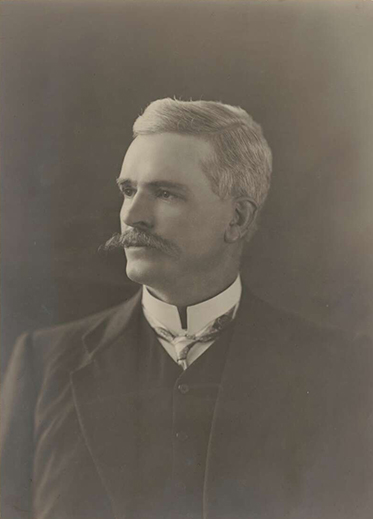 Faded, black and white studio portrait photo of silver-haired man with moustache.