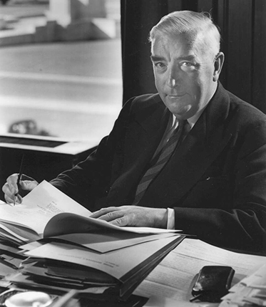 Menzies at his desk in Parliament House with piles of paper in front of him.