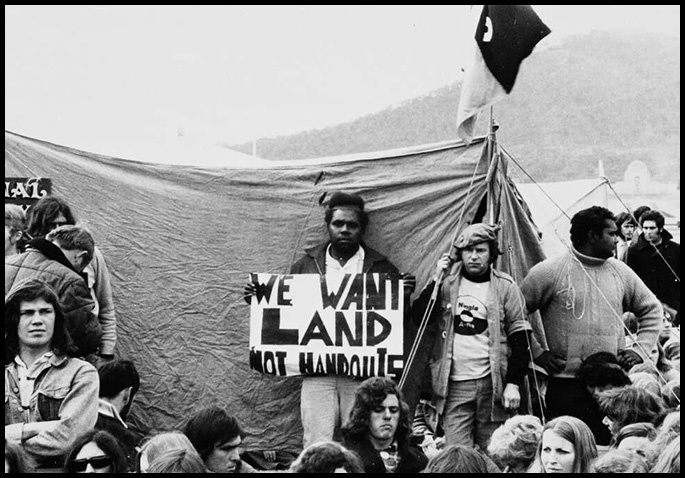 Group of young people, some of whom are Aboriginal, sitting or standing in front of a tent with a flag above it. An Aboriginal man at the centre of the photo is standing up looking at the camera holding a placard that says ‘We want land not handouts’.