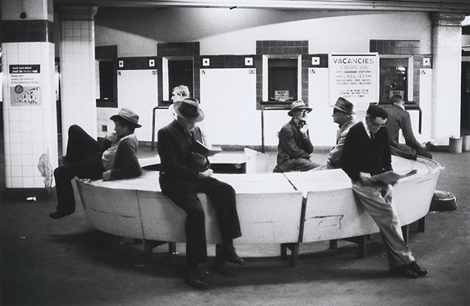 Interior photo of seven men sitting on a circular bench cum table. A few of them are reading but most are doing nothing at all. In the background are the station’s ticket booths. Next to one of these is a poster advertising railway jobs.