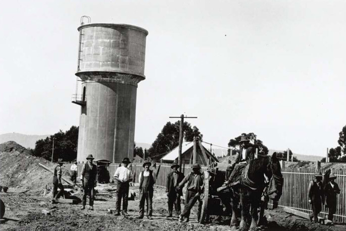 A dozen or so men at work on a suburban construction site, most of them posing for the camera. A draught horse attached to a cart stands among them eating from a nosebag. They appear to be clearing land. A water tower and suburban roofs are visible in the background