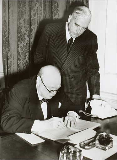Churchill is at a desk examining some documents. Menzies is standing beside him, looking over his shoulder