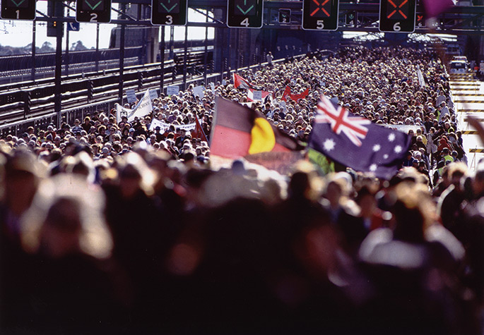 A vast crowd of people cover the roadway of the Bridge. People are carrying the Aboriginal and Australian flags.