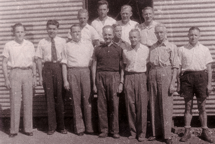 12 men, mostly wearing shirts and long pants, standing in front of a corrugated iron structure