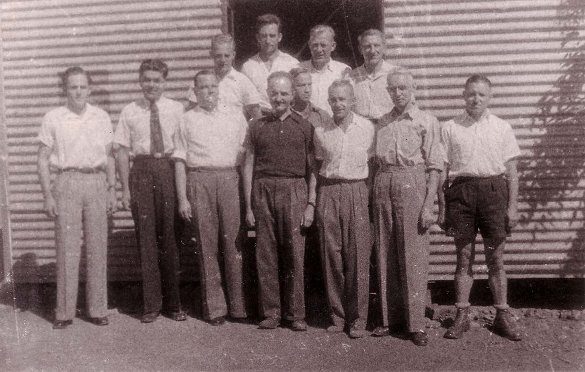 12 men, mostly wearing shirts and long pants, standing in front of a corrugated iron structure