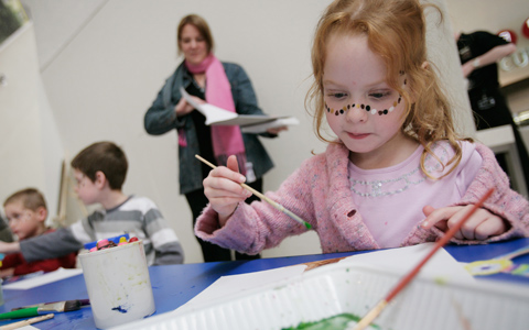 Three children at a table with crayons in a mug, paint brushes and paper. A girl, closest to the camera and on the right side of the image has dots painted across her face. An adult with additional paper is in the background, somewhat out of focus.