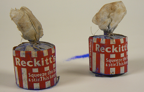 Two small off-white cylindrical cloth bags, tied at the top with string. Red and white striped paper is wrapped around each bag, with the text 'Reckitt's. ASqueeze the bag & stir. This keeps ...' Blue powder is visible on both bags.