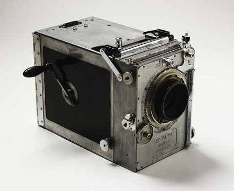 A rectangular box-shaped motion picture camera with a hand-crank and glass lens. The camera's metal body is silver with black side panels and has external slides, levers and dials for adjustment and operation. 'Le Parvo Modele L' is stamped in the metal plate beneath the lens at the front of the camera.