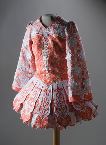 A peach-coloured dress with embroidery and applique in peach, white, yellow and gold. The dress has a detachable sash on the back. The skirt of the dress includes several petticoat layers. The top of the bodice and the sleeves are overlaid with white lace.