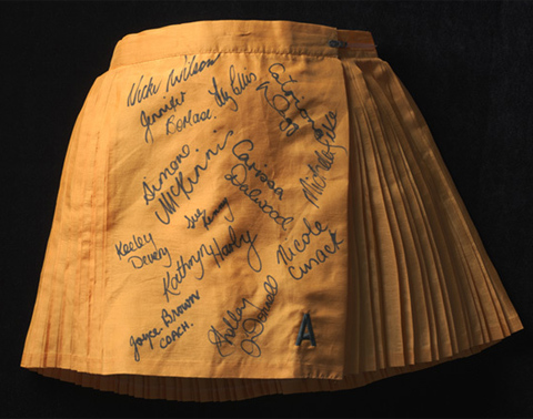 A golden yellow polyester netball skirt. The skirt is pleated, in alternating wide and narrow pleats. The front panel of the skirt is unpleated and signed in black ink by the members of the Australian women's netball team.
