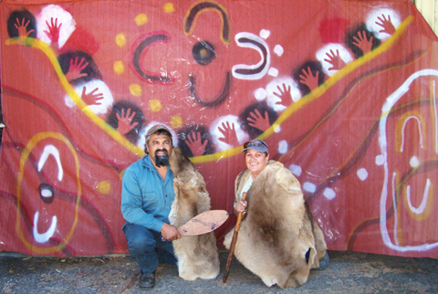 Man wearing a booka (kangaroo cloak) holding a spear thrower next to a woman also wearing a booka and holding a throwing stick