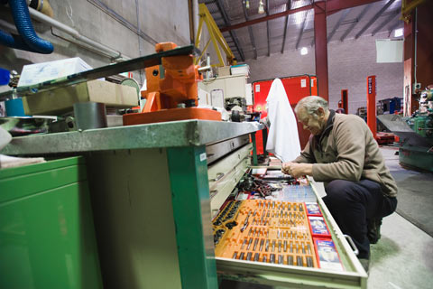 A man in an workshop kneels in front of an open drawer that contains various tools.