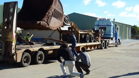 A view of a large mining excavator bucket being lifted off a semi-trailer. The semi-trailer faces diagonally left to right in the image, with its rear at the left-hand side of the image. The bucket has been lifted about a metre off the trailer. It faces toward the rear of the trailer. Two men are at either side of the trailer, retrieving long pieces of timber from the trailer's surface. Two more men are in the centre foreground of the image, with their backs to the camera. One squats on his haunches while the other is in a semi-kneeling position. Both are looking at the bucket. In the right-hand side of the image is an open concreted area. In the background of the image part of a large green industrial shed is visible. Beyond the shed can be seen some sky with a few clouds.