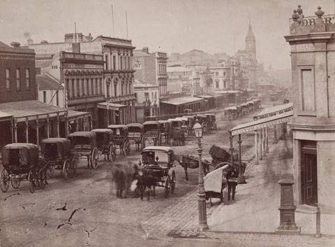 Black and white image of an urban streetscape with a series of two-storey buildings on either side. Many have verandahs at the front and a buildig with a spire rises towards the far end of the street. A series of horse-drawn carriages is lined up along the left side of the street. Another two carriages and several people stand to the right.